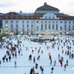 Eine große Gruppe von Menschen, die auf einer Freiluft-Eisbahn vor einem großen historischen Gebäude auf einem Stadtplatz Schlittschuh laufen.
