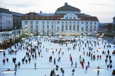 Eine große Gruppe von Menschen, die auf einer Freiluft-Eisbahn vor einem großen historischen Gebäude auf einem Stadtplatz Schlittschuh laufen.