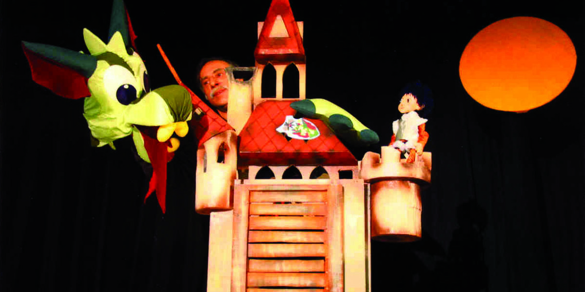 Eine Person bedient eine Drachenpuppe über einer Burgkulisse mit einer Puppenfigur auf einem Türmchen und einem orangefarbenen Kreis, der die Sonne im Hintergrund darstellt.