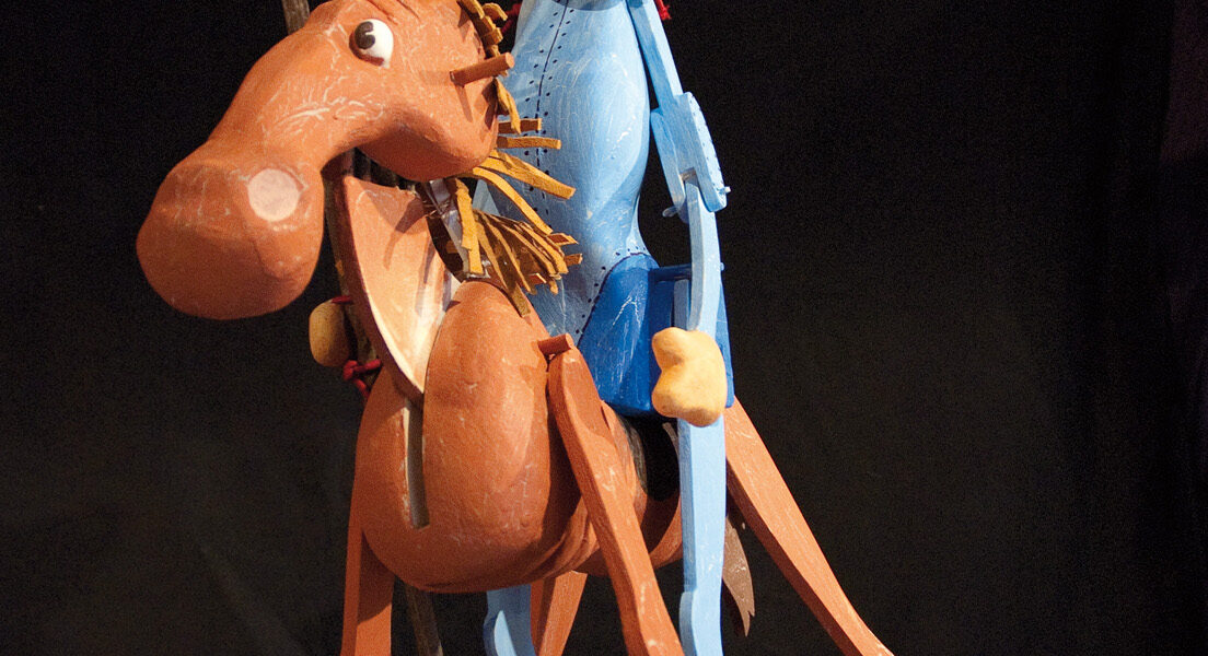 Eine hölzerne Puppenfigur eines Mannes mit Hut, der auf einem Pferd reitet und eine Lanze hält, vor einem dunklen Hintergrund.