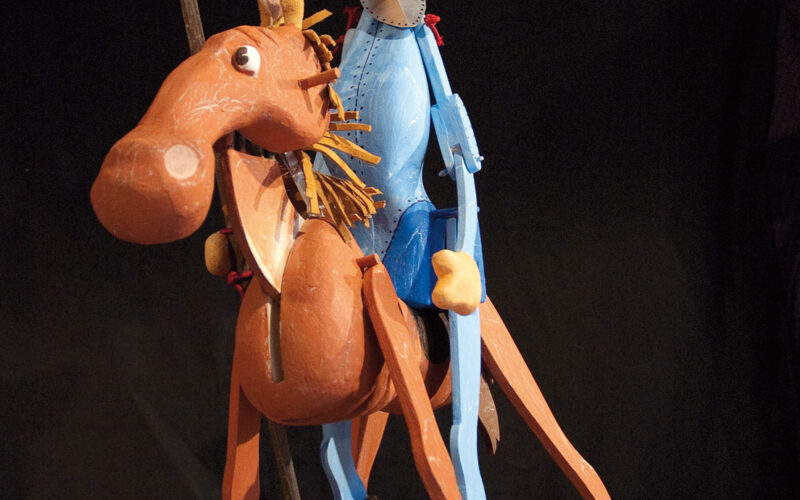 Eine hölzerne Puppenfigur eines Mannes mit Hut, der auf einem Pferd reitet und eine Lanze hält, vor einem dunklen Hintergrund.