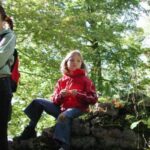 Ein Kind in einer roten Jacke sitzt auf einer niedrigen Steinmauer in einem Waldgebiet, während ein Erwachsener in der Nähe steht; das Sonnenlicht fällt durch die Bäume.