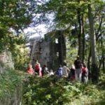 Eine Gruppe von Menschen steht in der Nähe der Ruinen eines alten Steingebäudes, umgeben von Bäumen und Grün.