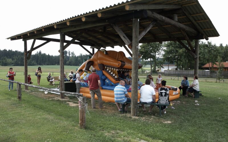 Die Menschen sitzen und stehen unter einem hölzernen Unterstand mit einer aufblasbaren Dinosaurierrutsche in einem grasbewachsenen Außenbereich.
