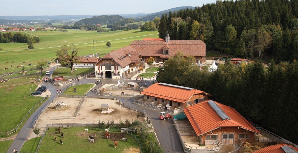 Luftaufnahme eines ländlichen Bauernhofs mit mehreren rotgedeckten Gebäuden, eingezäunten Tiergehegen, grünen Feldern und bewaldeten Flächen im Hintergrund.