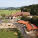 Luftaufnahme eines ländlichen Bauernhofs mit mehreren rotgedeckten Gebäuden, eingezäunten Tiergehegen, grünen Feldern und bewaldeten Flächen im Hintergrund.