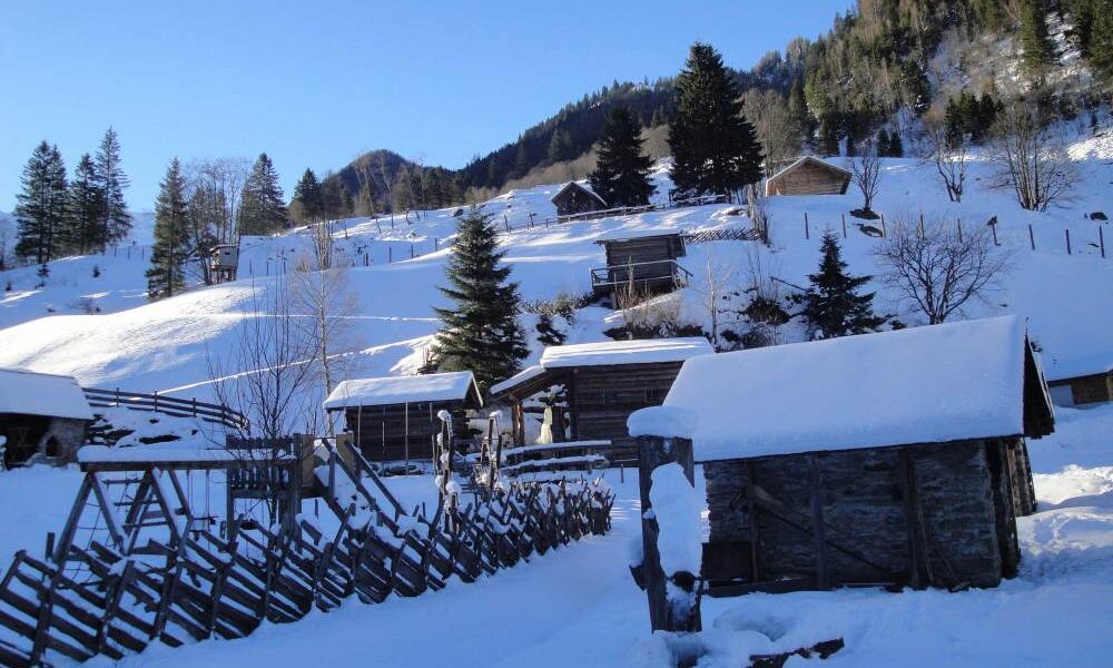 Schneebedeckte Holzhütten und Zäune an einem Hang, umgeben von Bäumen und Bergen unter einem klaren blauen Himmel.