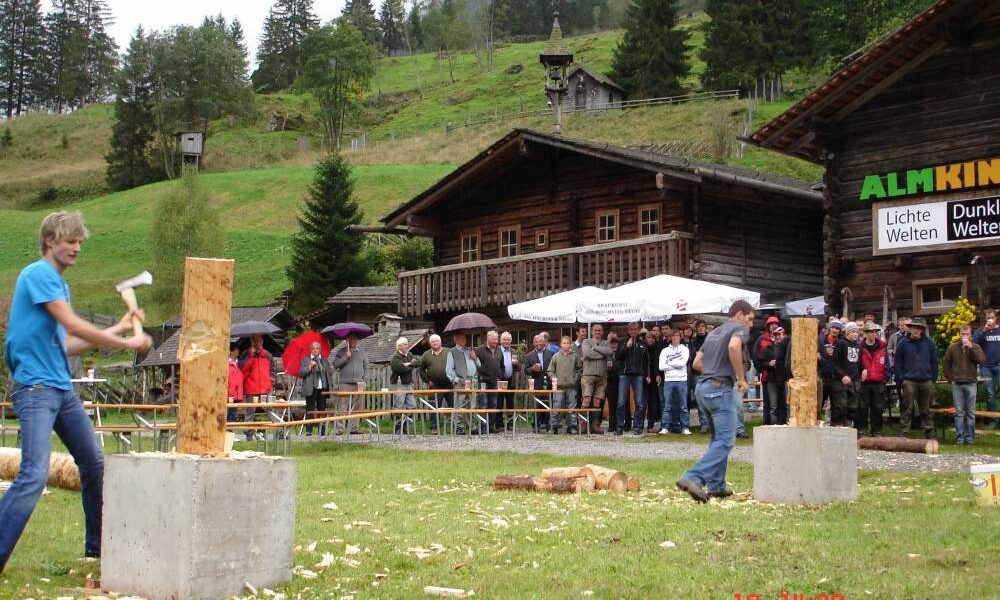 Zwei Personen nehmen an einem Holzhackwettbewerb im Freien teil, während eine Menschenmenge in der Nähe von Holzgebäuden und Bäumen im Hintergrund zusieht.