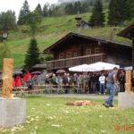 Zwei Personen nehmen an einem Holzhackwettbewerb im Freien teil, während eine Menschenmenge in der Nähe von Holzgebäuden und Bäumen im Hintergrund zusieht.