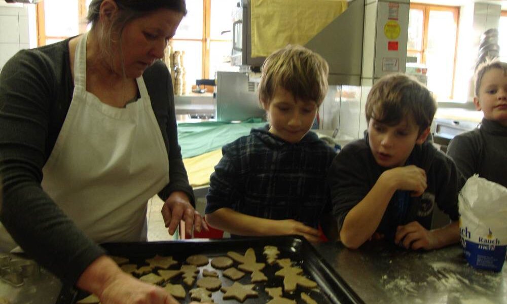 Eine Frau legt Plätzchenteigformen auf ein Backblech, während drei Kinder in einer Küche aufmerksam zusehen.