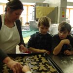 Eine Frau legt Plätzchenteigformen auf ein Backblech, während drei Kinder in einer Küche aufmerksam zusehen.