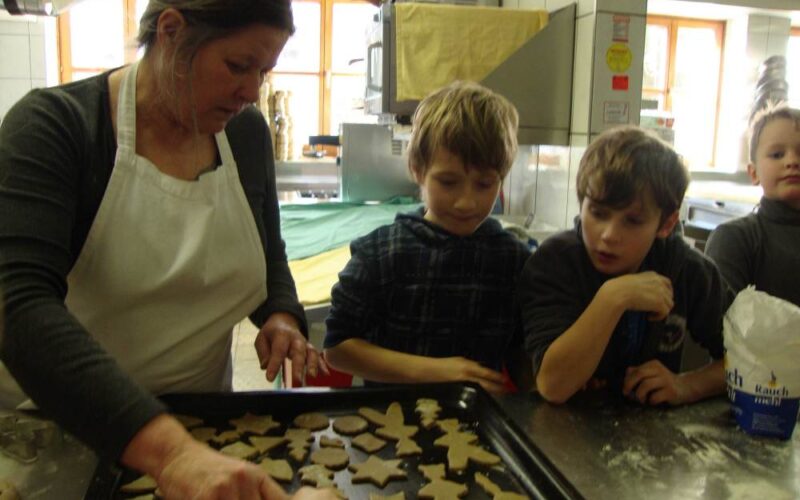 Eine Frau legt Plätzchenteigformen auf ein Backblech, während drei Kinder in einer Küche aufmerksam zusehen.