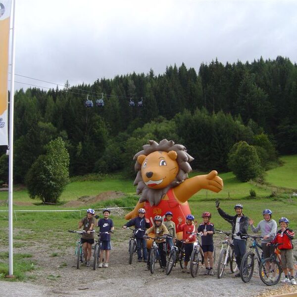 Eine Gruppe von Radfahrern posiert vor einem großen aufblasbaren Löwen im Freien, mit einem bewaldeten Berghang und Seilbahnen im Hintergrund.