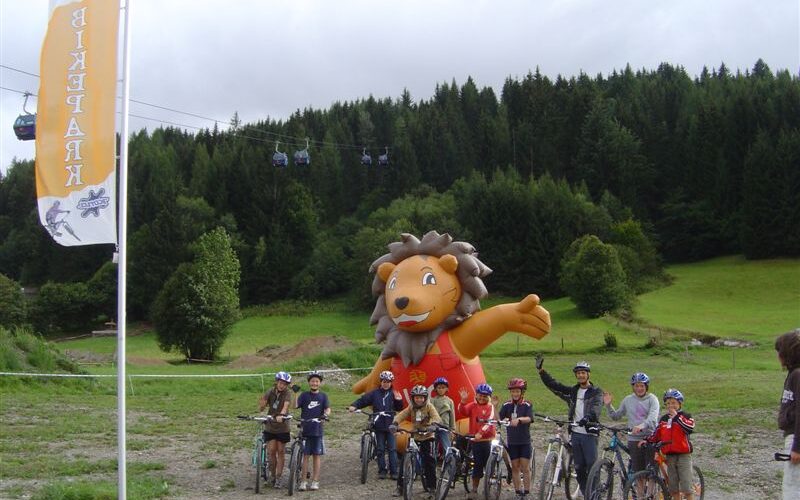 Eine Gruppe von Radfahrern posiert vor einem großen aufblasbaren Löwen im Freien, mit einem bewaldeten Berghang und Seilbahnen im Hintergrund.