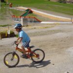 Ein Kind mit Helm fährt mit dem Fahrrad auf einem Kiesweg in der Nähe einer Rutsche und eines Grashügels, im Hintergrund ist ein gelbes Absperrband zu sehen.