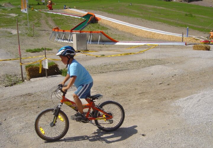 Ein Kind mit Helm fährt mit dem Fahrrad auf einem Kiesweg in der Nähe einer Rutsche und eines Grashügels, im Hintergrund ist ein gelbes Absperrband zu sehen.