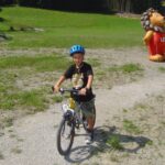 Ein lächelndes Kind, das einen blauen Helm trägt, fährt mit dem Fahrrad auf einem Kiesweg, mit einer großen aufblasbaren Löwenfigur und einer grasbewachsenen Wiese im Hintergrund.