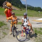 Zwei Kinder mit Helmen sitzen auf Fahrrädern auf einem Schotterweg mit einem großen aufblasbaren Löwen und grasbewachsenen Hügeln im Hintergrund.