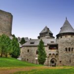 Eine steinerne Burg mit mehreren Türmen und kegelförmigen Dächern, umgeben von Bäumen und grünem Gras, unter einem klaren Himmel.