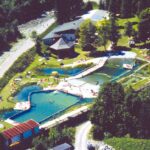 Luftaufnahme eines Freibadkomplexes mit mehreren Becken, Rasenflächen und Bäumen in der Nähe, neben einem Fluss und mehreren Gebäuden.