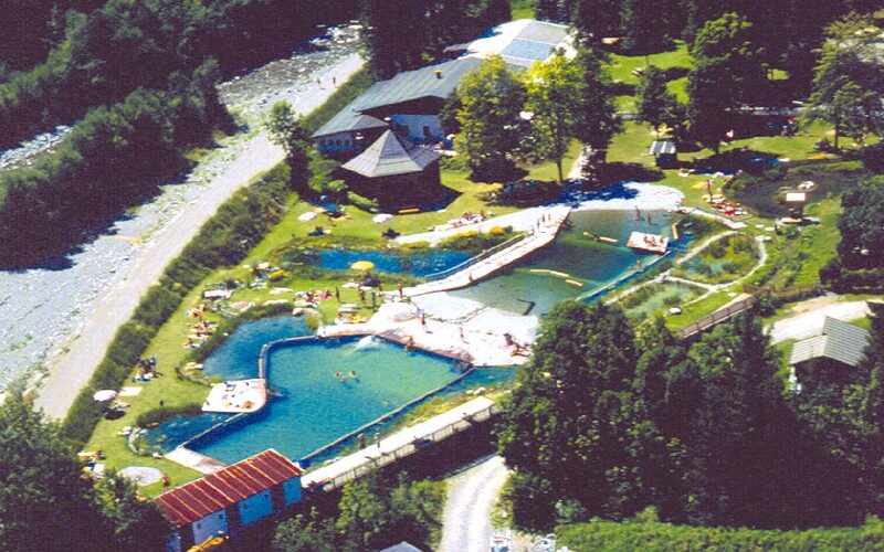 Luftaufnahme eines Freibadkomplexes mit mehreren Becken, Rasenflächen und Bäumen in der Nähe, neben einem Fluss und mehreren Gebäuden.