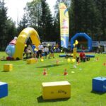 Kinderspielplatz im Freien mit aufblasbaren Strukturen, bunten Blöcken, Kegeln und Luftballons auf einer Wiese, wo Erwachsene und Kinder an Aktivitäten teilnehmen.