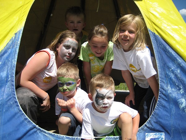 Sechs Kinder mit Gesichtsbemalung posieren an einem sonnigen Tag gemeinsam am Eingang eines gelb-blauen Zeltes.