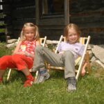 Zwei kleine Kinder sitzen in kleinen Liegestühlen auf einem grasbewachsenen Rasen vor einem Holzgebäude und lächeln in die Kamera.