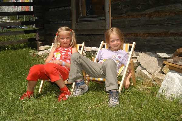 Zwei kleine Kinder sitzen in kleinen Liegestühlen auf einem grasbewachsenen Rasen vor einem Holzgebäude und lächeln in die Kamera.