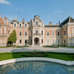 Ein großes, verziertes historisches Herrenhaus mit gemustertem Mauerwerk steht hinter einem gepflegten Garten und einem geschwungenen, spiegelnden Pool an einem klaren Tag.