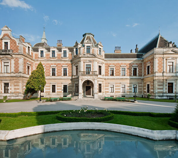 Ein großes, verziertes historisches Herrenhaus mit gemustertem Mauerwerk steht hinter einem gepflegten Garten und einem geschwungenen, spiegelnden Pool an einem klaren Tag.