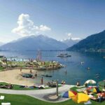 Die Menschen entspannen sich auf Gras- und Sandflächen an einem See mit Bergen im Hintergrund; einige schwimmen oder fahren mit Tretbooten, während andere sich unter Sonnenschirmen sonnen.