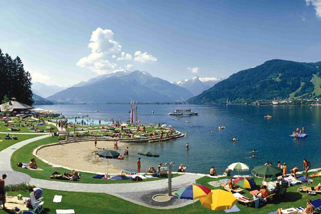 Die Menschen entspannen sich auf Gras- und Sandflächen an einem See mit Bergen im Hintergrund; einige schwimmen oder fahren mit Tretbooten, während andere sich unter Sonnenschirmen sonnen.