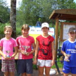 Vier Kinder stehen im Freien und halten Trophäen, im Hintergrund sind Informationstafeln und Grünpflanzen zu sehen.