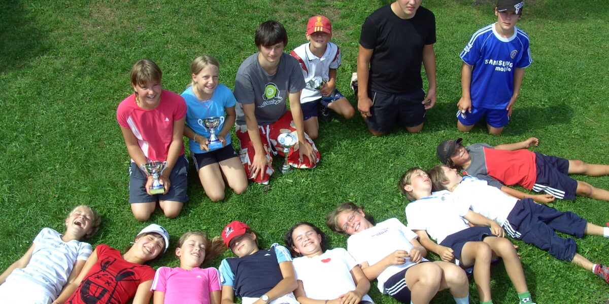 Eine Gruppe von Kindern posiert auf einer Wiese; einige stehen oder knien mit Trophäen, andere liegen lächelnd nebeneinander, in legerer Sportkleidung.