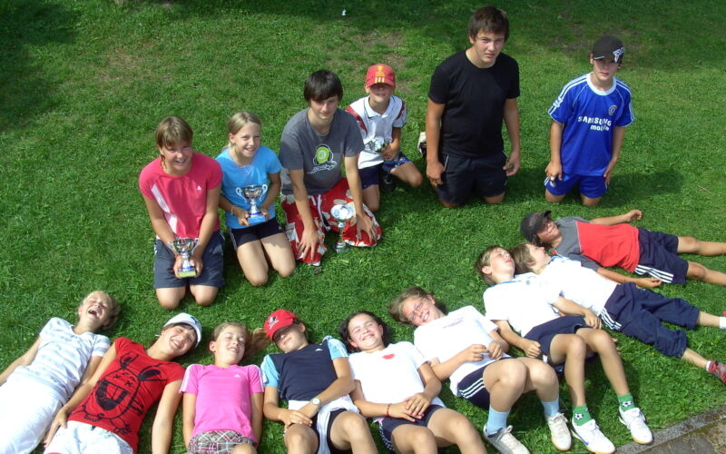Eine Gruppe von Kindern posiert auf einer Wiese; einige stehen oder knien mit Trophäen, andere liegen lächelnd nebeneinander, in legerer Sportkleidung.