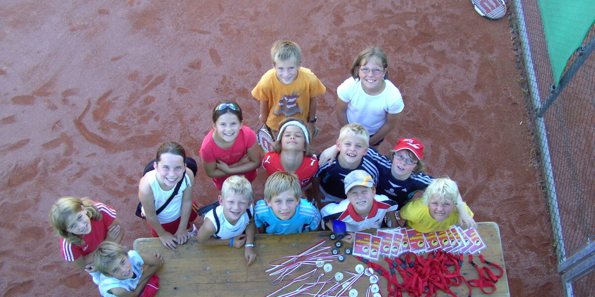 Eine Gruppe von Kindern steht um einen Tisch mit Medaillen und Bändern auf einem Tennisplatz, schaut auf und lächelt in die Kamera.