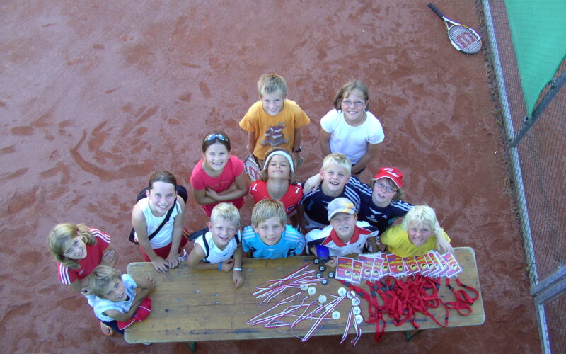 Eine Gruppe von Kindern steht um einen Tisch mit Medaillen und Bändern auf einem Tennisplatz, schaut auf und lächelt in die Kamera.