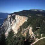 Ein felsiger, mit dichten Kiefern bewachsener Bergfelsen überragt ein Tal unter strahlend blauem Himmel. Ein gewundener Pfad führt durch den Wald am Rand entlang.