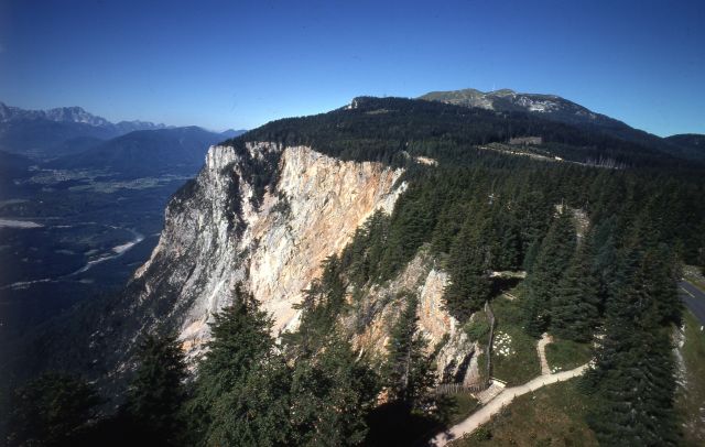 Ein felsiger, mit dichten Kiefern bewachsener Bergfelsen überragt ein Tal unter strahlend blauem Himmel. Ein gewundener Pfad führt durch den Wald am Rand entlang.