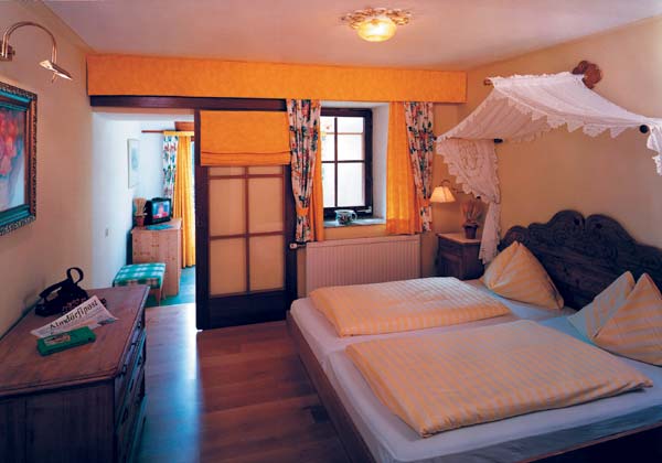 Ein gemütliches Schlafzimmer mit einem Doppelbett mit gelb gestreifter Bettwäsche, einem Kopfteil aus Holz, einem Spitzendach, einer Kommode, einem Fenster mit geblümten Vorhängen und warmer Beleuchtung.