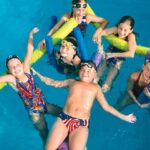 Sechs Kinder in Badeanzügen und Schwimmbrillen lächeln und spielen mit Poolnudeln in einem Schwimmbad, von oben gesehen.