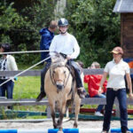 Eine Person mit einem Helm reitet auf einem Pferd über eine blau-weiße Stange, während eine andere Person mit einer Mütze daneben steht. Im Hintergrund stehen mehrere Personen in der Nähe eines Holzzauns.