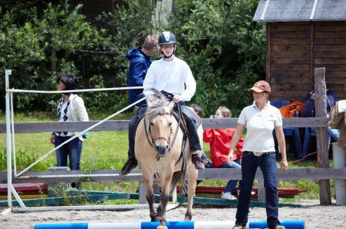 Eine Person mit einem Helm reitet auf einem Pferd über eine blau-weiße Stange, während eine andere Person mit einer Mütze daneben steht. Im Hintergrund stehen mehrere Personen in der Nähe eines Holzzauns.