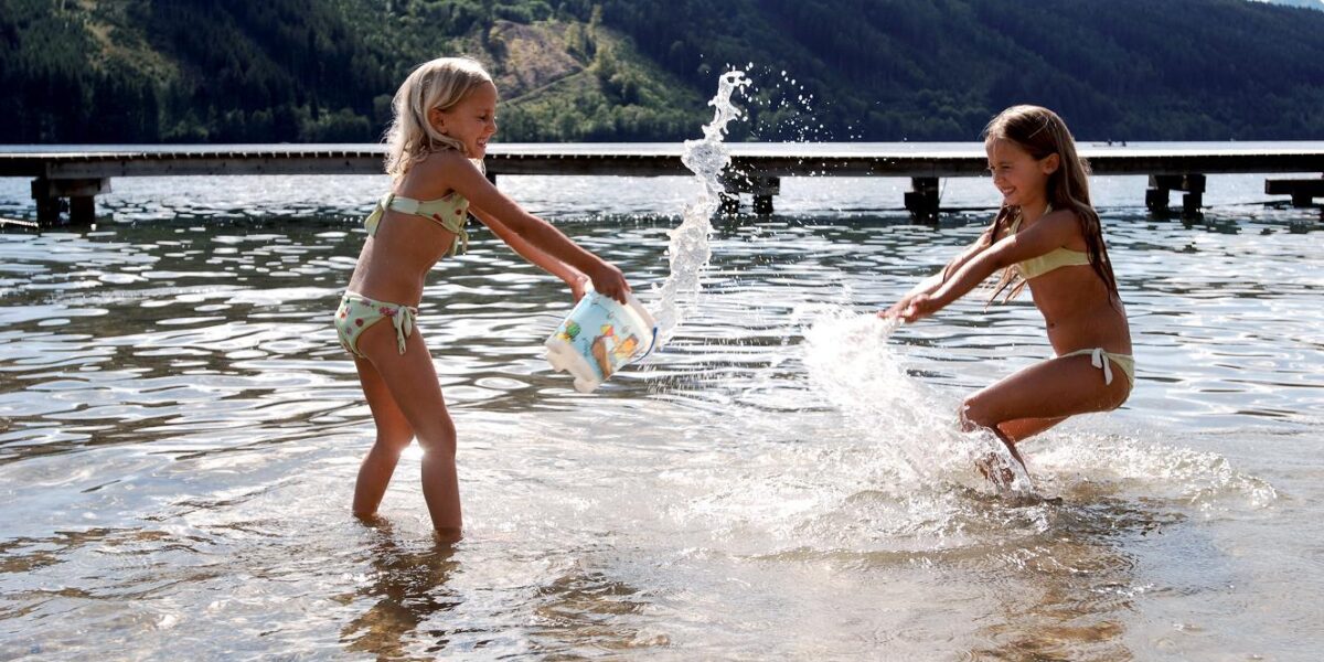 Zwei Mädchen in Badeanzügen spielen und bespritzen sich gegenseitig mit Wasser in einem flachen See, mit einem Steg und bewaldeten Bergen im Hintergrund.