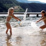 Zwei Mädchen in Badeanzügen spielen und bespritzen sich gegenseitig mit Wasser in einem flachen See, mit einem Steg und bewaldeten Bergen im Hintergrund.