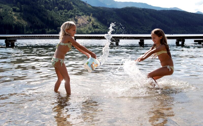 Zwei Mädchen in Badeanzügen spielen und bespritzen sich gegenseitig mit Wasser in einem flachen See, mit einem Steg und bewaldeten Bergen im Hintergrund.