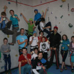 Eine Gruppe von Kindern und Erwachsenen posiert vor einer Indoor-Kletterwand, einige tragen Klettergurte und Helme, halten Karten und Kletterausrüstung.