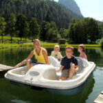 Eine Frau und drei Kinder sitzen in einem weißen Paddelboot auf einem ruhigen See, der an einem sonnigen Tag von Bäumen und Bergen umgeben ist.