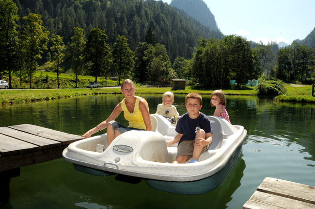 Eine Frau und drei Kinder sitzen in einem weißen Paddelboot auf einem ruhigen See, der an einem sonnigen Tag von Bäumen und Bergen umgeben ist.